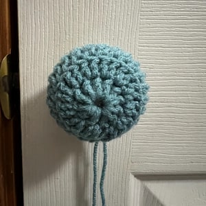 Puede incluir: Un cubre pomo de puerta de crochet azul claro con una cuerda larga que cuelga.
