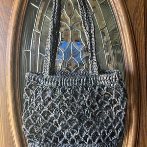 May include: A dark gray woven tote bag with braided handles. The bag has an open, net-like design. The bag is hanging on a wooden door with a glass window.
