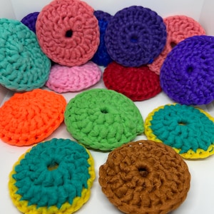 May include: A collection of colourful crocheted scrubbers in various colours, including pink, purple, green, orange, brown, and teal. The scrubbers are all round and have a hole in the centre.