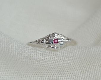 May include: A silver ring featuring a textured, organic-shaped band with a small, round, vibrant red gemstone set in the center. The metal surface has an uneven, molten appearance with subtle indentations and raised areas across the band.
