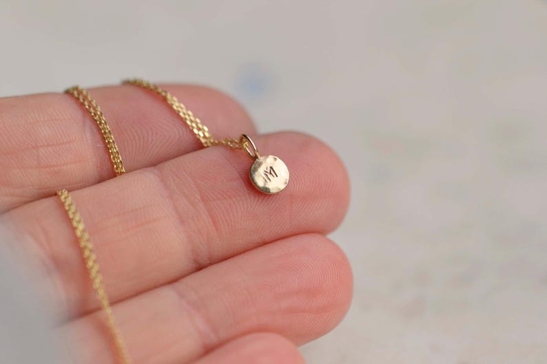 Pu&ograve; includere: Una collana color oro con una catenina sottile e un piccolo ciondolo rotondo dalla texture martellata. Il ciondolo presenta la lettera maiuscola M impressa al centro. Il metallo ha una finitura lucida e una calda tonalit&agrave; giallo metallico.
