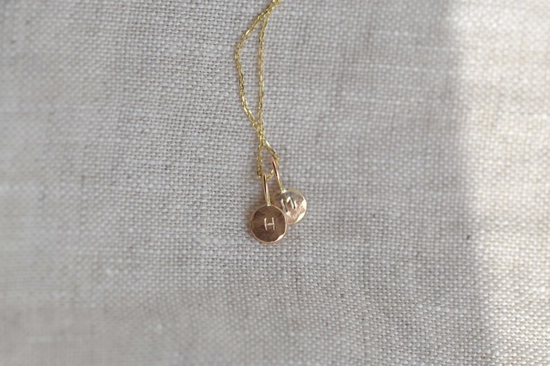 Pu&ograve; includere: Una collana color oro con due piccoli ciondoli circolari e piatti sospesi a una catenina sottile. Ogni ciondolo &egrave; stampato con una singola lettera maiuscola, uno mostra la lettera H e l'altro mostra la lettera M.