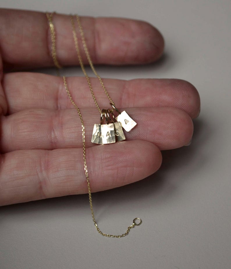 Pu&ograve; includere: Una collana a catena in oro con quattro piccoli ciondoli rettangolari in oro. Tre dei ciondoli sono stampati con le lettere M, A e C, mentre il quarto ciondolo &egrave; stampato con la lettera A. I ciondoli pendono da una sottile catena a maglie d'oro.