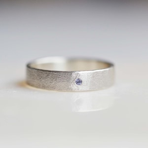 May include: A silver ring with a textured finish and a small, round, pale purple gemstone. The ring is set against a white background. The ring's details are in soft focus.