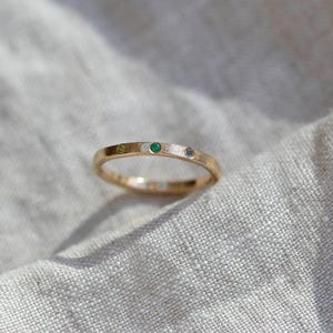 Puede incluir: Un anillo de banda fina de oro con tres peque&ntilde;as piedras preciosas engastadas al ras en fila. Las piedras incluyen una gema verde claro, una esmeralda verde vibrante y un diamante blanco claro, incrustadas en la superficie met&aacute;lica texturizada.