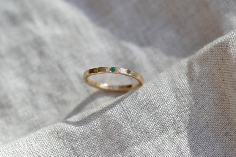 Puede incluir: Un anillo de banda fina de oro con tres peque&ntilde;as piedras preciosas engastadas al ras en fila. Las piedras incluyen una gema verde claro, una esmeralda verde vibrante y un diamante blanco claro, incrustadas en la superficie met&aacute;lica texturizada.
