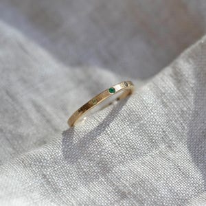 Puede incluir: Un anillo de banda fina de oro que presenta una peque&ntilde;a piedra preciosa verde redonda engastada en la superficie met&aacute;lica. El anillo tiene un acabado liso y pulido, con un perfil sencillo y delgado que mantiene un ancho constante en toda la banda.
