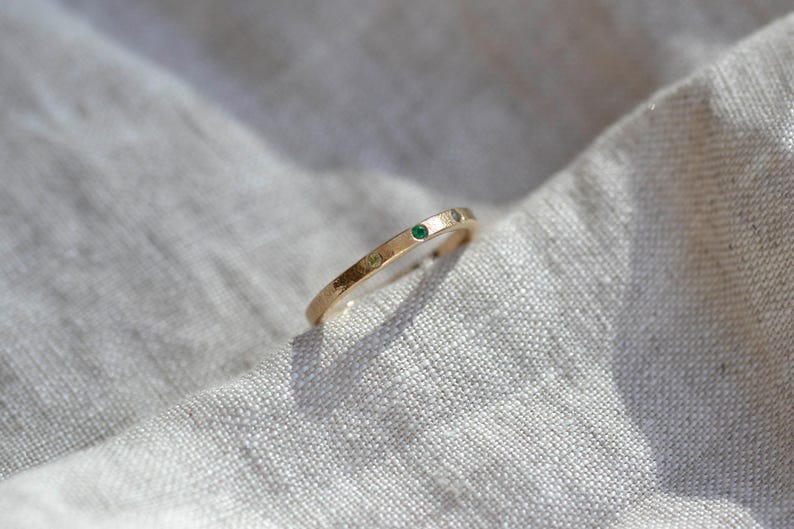 Puede incluir: Un anillo de banda fina de oro que presenta una peque&ntilde;a piedra preciosa verde redonda engastada en la superficie met&aacute;lica. El anillo tiene un acabado liso y pulido, con un perfil sencillo y delgado que mantiene un ancho constante en toda la banda.