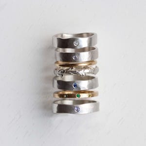 Puede incluir: Una pila vertical de siete anillos con diversos dise&ntilde;os, incluyendo bandas de plata lisas con piedras incrustadas, una banda de plata texturizada, una banda de oro lisa y una banda de oro con tres peque&ntilde;as piedras verdes en fila.
