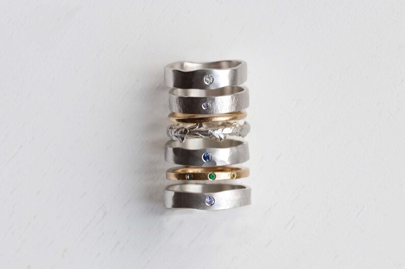 Puede incluir: Una pila vertical de siete anillos con diversos dise&ntilde;os, incluyendo bandas de plata lisas con piedras incrustadas, una banda de plata texturizada, una banda de oro lisa y una banda de oro con tres peque&ntilde;as piedras verdes en fila.