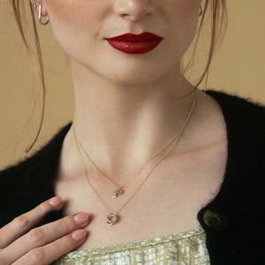 Pu&ograve; includere: Una collana a strati in oro composta da due catenine sottili. La catenina pi&ugrave; corta presenta due piccoli ciondoli rettangolari, mentre quella pi&ugrave; lunga ha un pendente circolare formato da tre anelli intrecciati di diverse tonalit&agrave; metalliche.