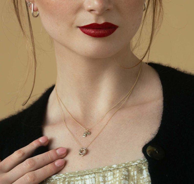Pu&ograve; includere: Una collana a strati in oro composta da due catenine sottili. La catenina pi&ugrave; corta presenta due piccoli ciondoli rettangolari, mentre quella pi&ugrave; lunga ha un pendente circolare formato da tre anelli intrecciati di diverse tonalit&agrave; metalliche.