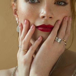 May include: A woman with red lipstick and freckles is holding her hands to her face. She is wearing a gold ring on her left ring finger and a silver ring on her right ring finger. She is also wearing a silver necklace and a pair of silver earrings.