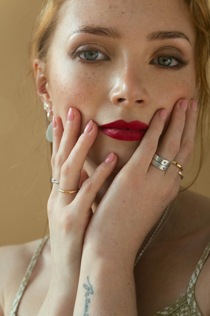 May include: A woman with red lipstick and freckles is holding her hands to her face. She is wearing a gold ring on her left ring finger and a silver ring on her right ring finger. She is also wearing a silver necklace and a pair of silver earrings.