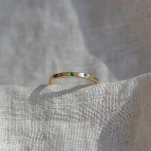 Puede incluir: Un anillo de banda fina de oro con tres peque&ntilde;as piedras preciosas engastadas al ras en la superficie superior. Las piedras incluyen una gema amarilla a la izquierda, una gema verde vibrante en el centro y una gema clara e incolora a la derecha.