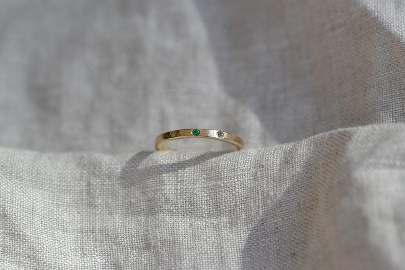 Puede incluir: Un anillo de banda fina de oro con tres peque&ntilde;as piedras preciosas engastadas al ras en la superficie superior. Las piedras incluyen una gema amarilla a la izquierda, una gema verde vibrante en el centro y una gema clara e incolora a la derecha.