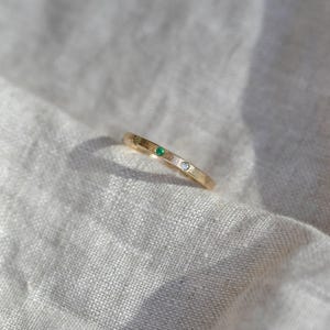 Puede incluir: Un anillo de banda fina de oro con dos peque&ntilde;as piedras preciosas engastadas al ras. Una piedra es de un verde vibrante y la otra es una piedra clara e incolora. El anillo tiene un acabado pulido y liso con un perfil sencillo y delgado.