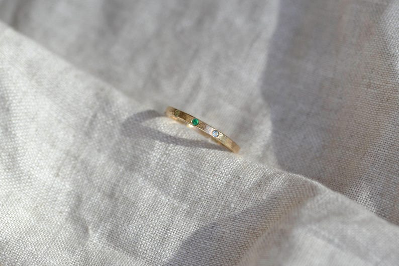 Puede incluir: Un anillo de banda fina de oro con dos peque&ntilde;as piedras preciosas engastadas al ras. Una piedra es de un verde vibrante y la otra es una piedra clara e incolora. El anillo tiene un acabado pulido y liso con un perfil sencillo y delgado.