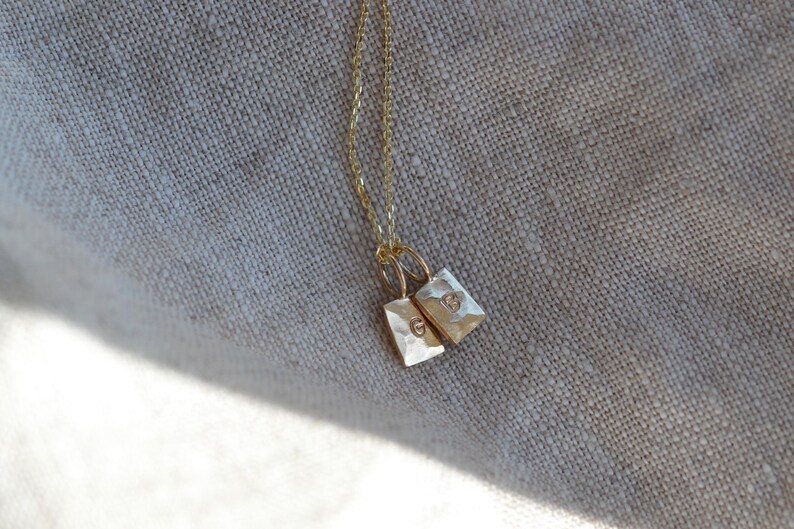 Pu&ograve; includere: Una collana a catena dorata con due piccoli ciondoli rettangolari dalla texture martellata. Ogni ciondolo &egrave; sospeso a un anellino e presenta una singola lettera stampata, con una che mostra la lettera G e l'altra la lettera B.