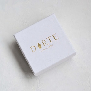 Pu&ograve; includere: Una scatola portagioie quadrata in cartone bianco con il testo in lamina d'oro DARTE JEWELLERY stampato sul coperchio. La lettera A nella parola DARTE &egrave; sostituita da una piccola icona a forma di albero triangolare dorato. Finitura opaca.