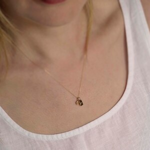 Pu&ograve; includere: Una delicata collana a catena dorata con un piccolo ciondolo rettangolare e un minuscolo ciondolo circolare in oro. La collana poggia su un top bianco senza maniche, mettendo in risalto un design essenziale con una sottile catena in metallo.