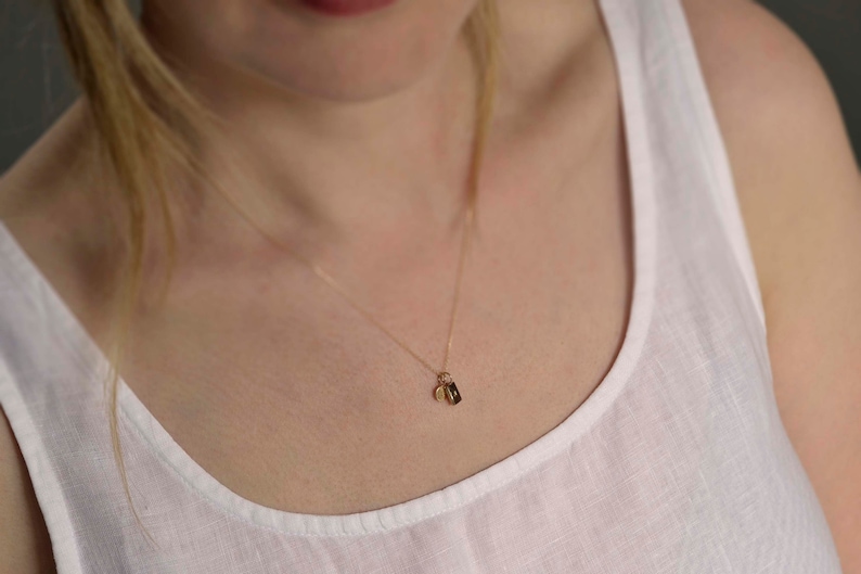 Pu&ograve; includere: Una delicata collana a catena dorata con un piccolo ciondolo rettangolare e un minuscolo ciondolo circolare in oro. La collana poggia su un top bianco senza maniche, mettendo in risalto un design essenziale con una sottile catena in metallo.