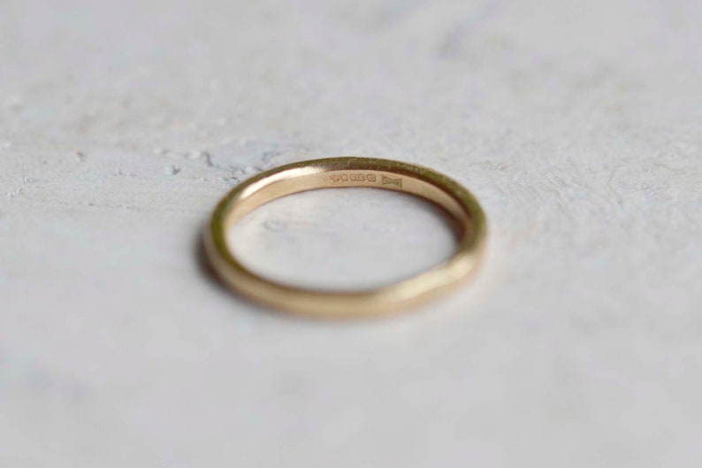 Puede incluir: Un anillo de banda de oro liso con un perfil redondeado. La superficie interior del anillo presenta una serie de peque&ntilde;os sellos grabados que son visibles tras una inspecci&oacute;n detallada, indicando la pureza del metal y su origen.