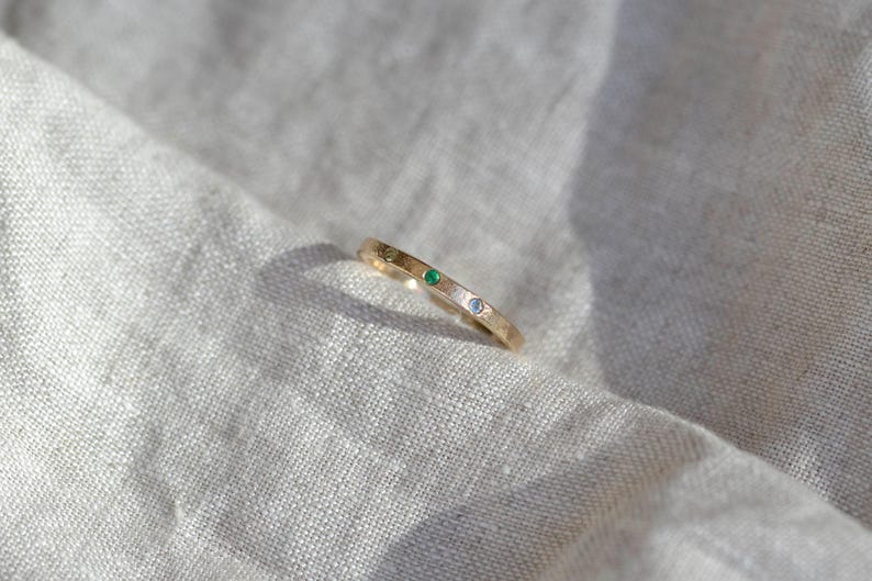 Puede incluir: Un anillo de banda fina de oro con tres peque&ntilde;as piedras preciosas engastadas al ras. Las piedras est&aacute;n dispuestas en fila en la parte superior de la banda, consistiendo en una piedra amarilla, una verde vibrante y una azul claro, incrustadas en el metal.