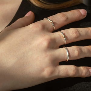 May include: Three silver rings with gold accents. The ring on the index finger has a small gold ball on the top. The ring on the middle finger has two gold beads. The ring on the little finger is a simple band.