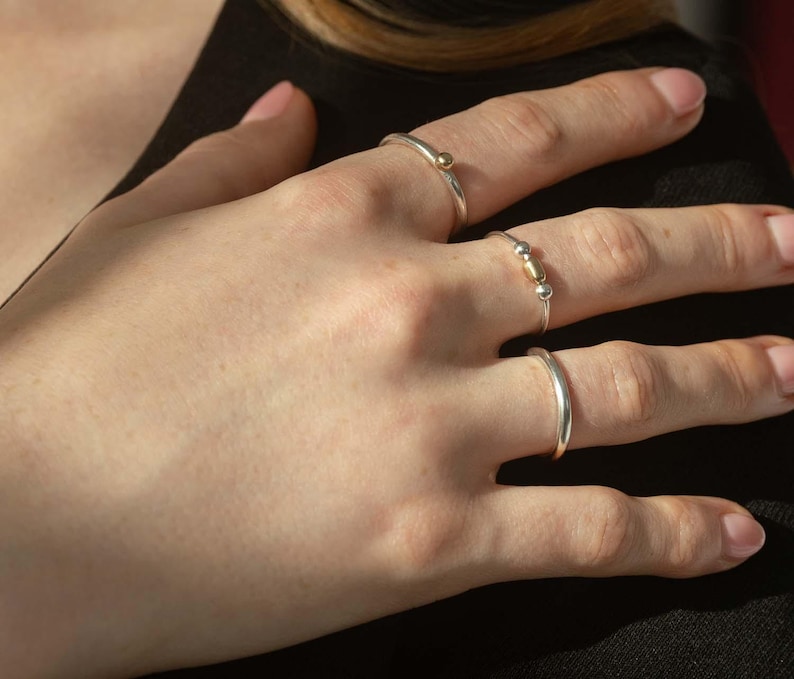 May include: Three silver rings with gold accents. The ring on the index finger has a small gold ball on the top. The ring on the middle finger has two gold beads. The ring on the little finger is a simple band.