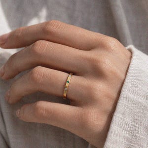 Puede incluir: Un anillo de banda fina de oro con una piedra preciosa verde central flanqueada por dos piedras blancas transparentes. La superficie del metal tiene un acabado texturizado sutil y las piedras est&aacute;n engastadas al ras en la banda, creando un perfil uniforme.
