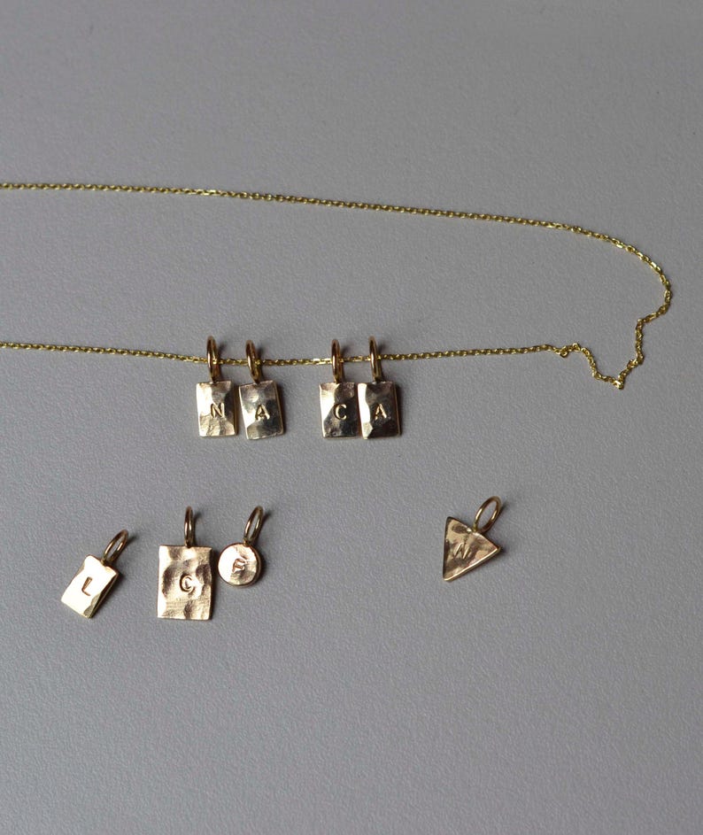 Pu&ograve; includere: Una collana a catena dorata con quattro ciondoli rettangolari stampati con le lettere N, A, C e A. Sotto la collana ci sono quattro ciondoli sciolti: un piccolo rettangolo con L, un rettangolo grande con C, un cerchio piccolo con E e un triangolo con M.