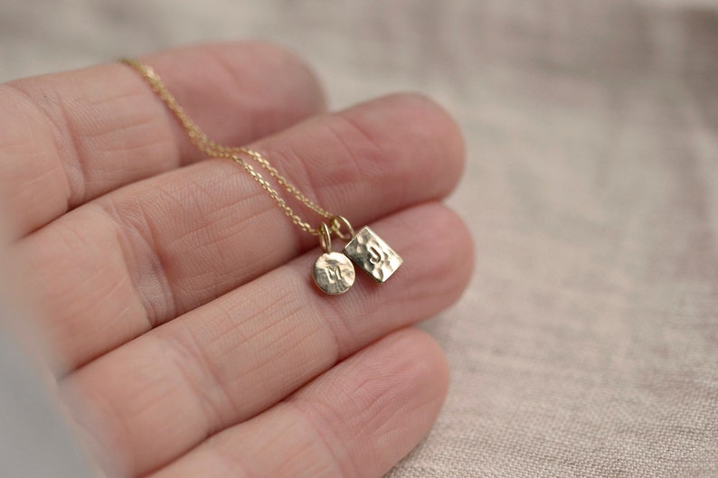 Pu&ograve; includere: Una collana color oro con due piccoli ciondoli in metallo martellato sospesi a una catenina sottile. Un ciondolo &egrave; circolare e l'altro rettangolare, entrambi presentano una lettera stampata sulle loro superfici testurizzate e riflettenti.