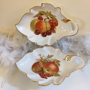 May include: Two white, leaf-shaped porcelain serving dishes with gold trim. Each dish features a hand-painted still life of fruit, including apples, cherries, and currants. The dishes are displayed on a white surface.