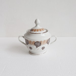 May include: A white ceramic sugar bowl with a lid, decorated with brown and tan floral patterns. The bowl has two handles and a decorative finial. A classic piece for serving sugar.