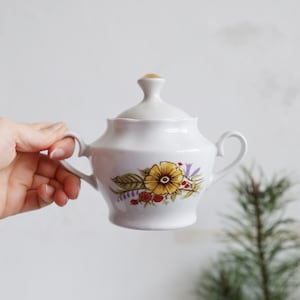 Könnte beinhalten: Weißer Keramik-Zuckertopf mit Deckel und zwei Griffen. Die Schale ist mit einem Blumendesign mit gelben und orangenen Blumen, grünen Blättern und lila Akzenten verziert. Ein kleiner, goldfarbener Knopf sitzt auf dem Deckel.