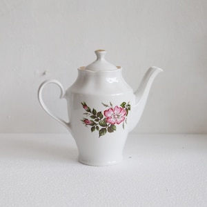 Puede incluir: Tetera de porcelana blanca con un diseño floral de rosas rosas y hojas verdes. La tetera tiene un asa curva, un pico y una tapa con un pequeño pomo. Un diseño clásico, adecuado para servir té.