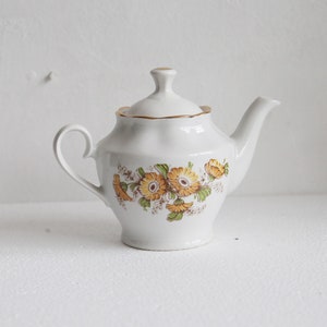 May include: White ceramic teapot with a lid, featuring a floral design in yellow, orange, and green. The teapot has a curved handle and a spout. Gold trim accents the lid and rim. A classic tea accessory.