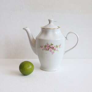 Puede incluir: Tetera de porcelana blanca con borde dorado y diseño floral de rosas rosas. La tetera tiene un asa curva y un pico largo. Una lima verde está al lado de la tetera.