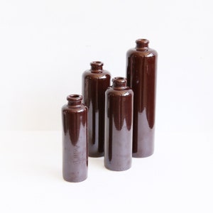 May include: Four brown ceramic bottles of varying heights. The bottles have a glossy finish and a narrow neck. They are arranged on a white surface against a white background. The bottles are cylindrical in shape.