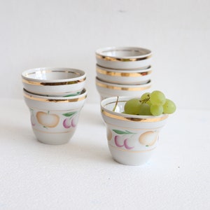 May include: A collection of small, white ceramic bowls with gold rims and fruit designs. One bowl holds a cluster of green grapes. The bowls are stacked in groups, showcasing their decorative patterns and elegant design.