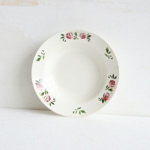 May include: A white ceramic plate with a shallow bowl shape. The rim is decorated with pink roses and green leaves. The plate is set against a white background, highlighting the floral design.