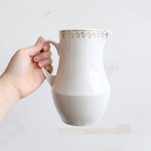 May include: A white ceramic pitcher with a gold decorative border around the rim. The bottom half of the pitcher is a light gray color. The pitcher has a handle and a spout for pouring.