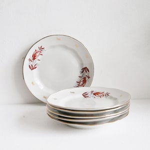 May include: A stack of four white ceramic plates with a gold rim and a floral design in red and brown. Another plate is standing upright behind the stack. The plates are on a white surface.