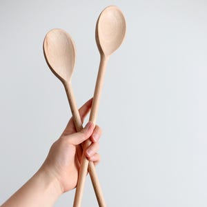 Può includere: Due cucchiai di legno con manici lunghi sono tenuti in una mano su sfondo bianco. I cucchiai sono fatti di legno chiaro e hanno delle conche di forma ovale. I manici sono sottili e cilindrici.