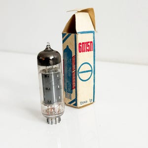 Puede incluir: Una válvula de vacío vintage con tapa negra y cuerpo de vidrio transparente, junto a su caja de cartón original. La caja es beige con gráficos rojos y azules y el texto "6M15П".