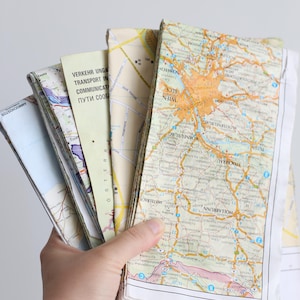 May include: A hand holds a stack of vintage travel maps, showing detailed road networks and city outlines. The maps display a variety of colours, with text in multiple languages. The maps are folded and show signs of wear.