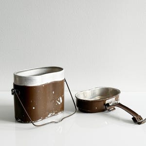 May include: A brown and silver metal mess kit consisting of a deep, oval-shaped container with a wire bail handle and a separate, shallower lid that features a folding metal handle. Both pieces show signs of wear with chipped brown paint.