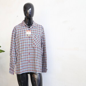 Peut inclure: Chemise à carreaux à manches longues, dans les tons bordeaux, bleu et blanc, présentée sur un mannequin. La chemise a une fermeture boutonnée, une poche poitrine et un col. Une étiquette de prix est attachée.