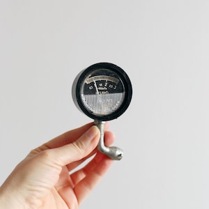 Può includere: Un manometro nero vintage con un manico argentato. Il manometro ha un quadrante circolare con un ago e numeri da 0,5 a 3. Il testo "-100kPa" è visibile. Il manometro è tenuto in una mano.